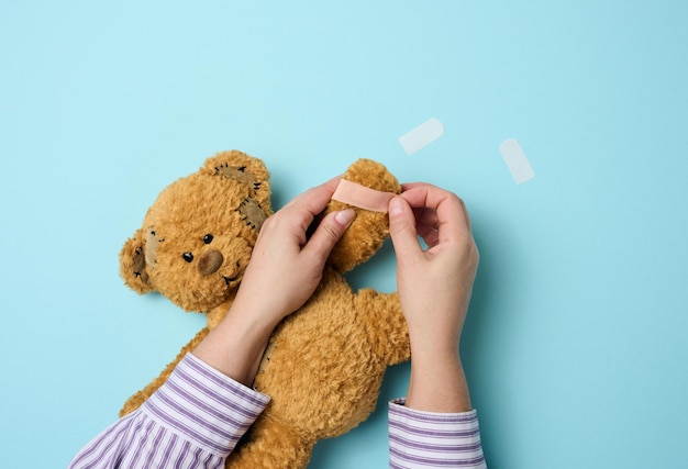 Mano femenina sostiene un oso de peluche marrón y pega un yeso adhesivo médico en un tratamiento de tranvía de fondo azul