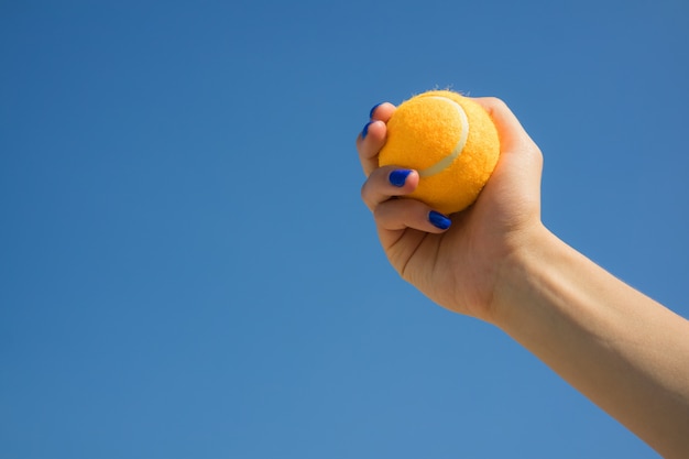 La mano femenina sostiene una pelota de tenis anaranjada brillante en un fondo del cielo azul