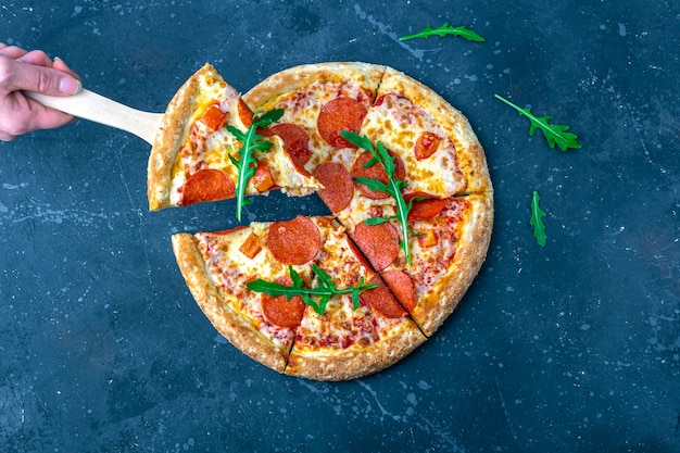 Mano femenina sostiene pizza con pala culinaria. Pizza de pepperoni recién preparada con salami y queso sobre un fondo oscuro. Almuerzo o cena tradicional italiana.