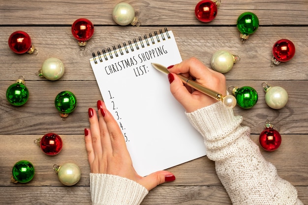 Mano femenina sostiene la pluma, escribiendo la lista de compras de Navidad, ideas para regalos en el Bloc de notas blanco sobre fondo de madera