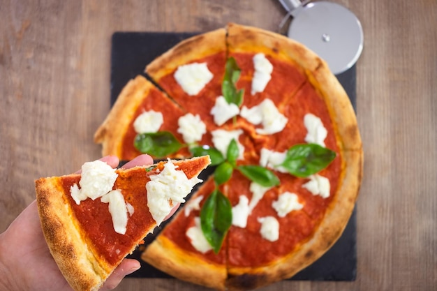 La mano femenina sostiene una rebanada de pizza italiana Margherita con pizza de tomate y mozzarella de búfala cubierta con albahaca fresca en el fondo de la pizarra negra