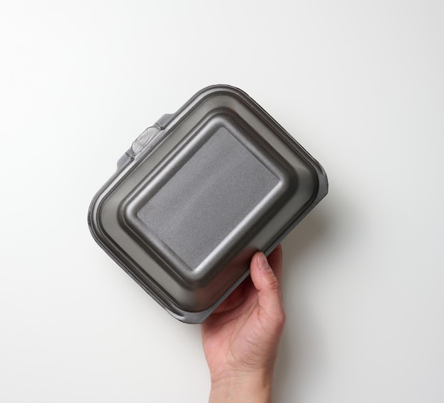 La mano femenina sostiene un recipiente de comida desechable. Caja gris de poliestireno sobre un fondo blanco.