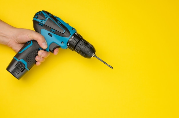 Mano femenina sostiene un taladro inalámbrico sobre un fondo amarillo. Herramienta para el trabajo