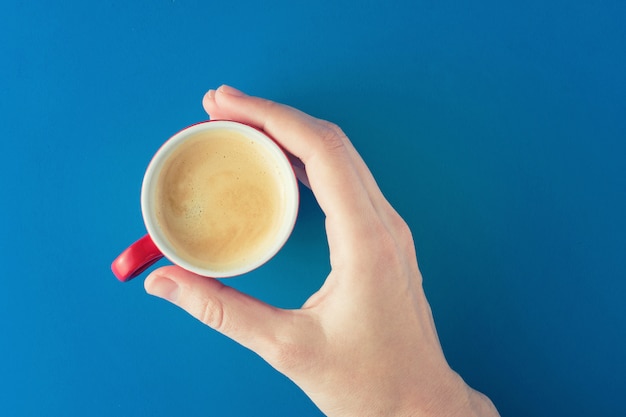 La mano femenina sostiene una taza de café roja en un fondo azul