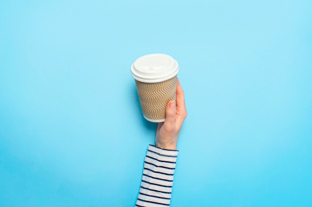 La mano femenina sostiene una taza de papel con café en un azul. Concepto de café, una bebida caliente, desayuno.