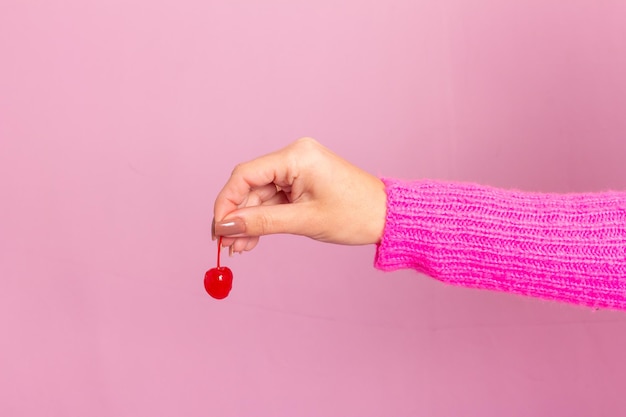 Foto mano femenina con suéter rosa y sosteniendo una cereza. fondo rosa claro.