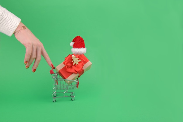 Mano femenina tirar de un carrito de supermercado carrito de compras con caja de regalo de Navidad vistiendo gorro de Papá Noel sobre un fondo verde