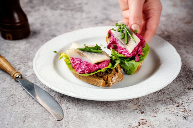 Mano femenina tomando sándwich con arenque, remolacha y ensalada verde de la placa