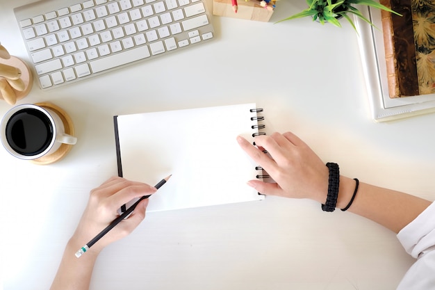 Mano femenina de la visión superior que escribe el cuaderno en blanco en el escritorio y el ordenador blancos de oficina con los materiales de oficina. concepto de espacio de trabajo