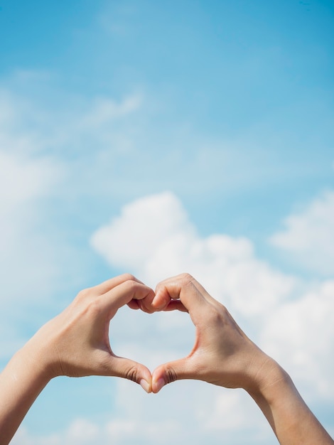 Foto mano en forma de corazón sobre cielo azul y fondo de nubes mullidas, concepto de amor, relación y estilo romántico, vertical. mano femenina haciendo signo de amor de dedo con espacio de copia.