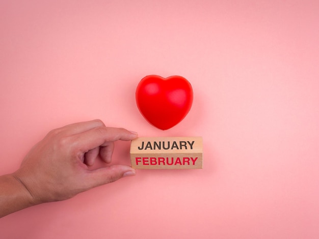 Mano girando un bloque de cubo de madera para cambiar el mes de enero a febrero y una bola de corazón rojo sobre fondo rosa pastel. Concepto de día de san valentín, amor, estilo minimalista.