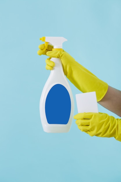 La mano en el guante amarillo contiene una botella de spray de detergente líquido y una esponja sobre fondo azul.