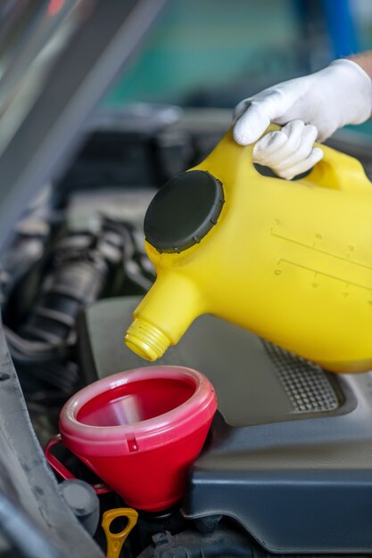 Mano en guante blanco protector sosteniendo un pequeño bote amarillo debajo del capó del coche