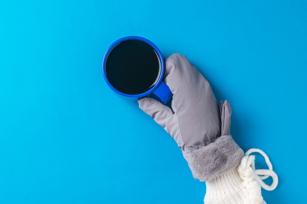 Una mano en un guante caliente con una taza de café caliente sobre una superficie azul. Bebida caliente y guantes.