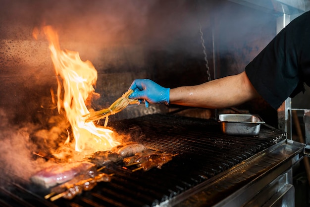 Mano con guante de un cocinero profesional cubriendo con especias un trozo de carne a la parrilla con una gran llama saliendo de la parrilla.