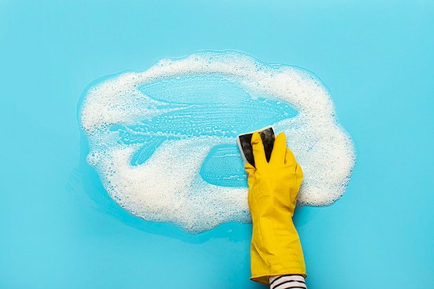 La mano en un guante de goma amarillo sostiene una esponja limpiadora y limpia una espuma jabonosa sobre una superficie azul. Concepto de limpieza, servicio de limpieza. . Vista plana, vista superior