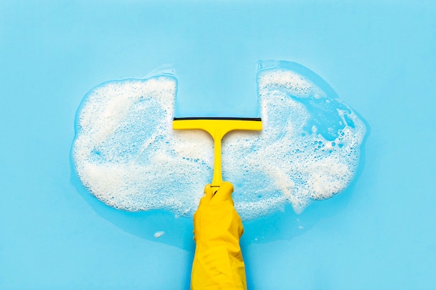 La mano en un guante de goma amarillo sostiene un raspador para limpiar y limpia la espuma de jabón sobre una superficie azul. Concepto de limpieza, servicio de limpieza. . Vista plana, vista superior