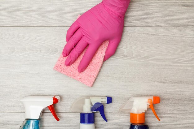 Mano en guante de goma sosteniendo una servilleta rosa con botellas de detergente en tablas de madera