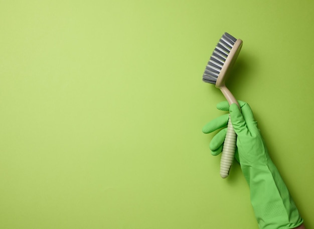 Mano en un guante de goma verde sostiene un cepillo de limpieza de plástico sobre un fondo verde
