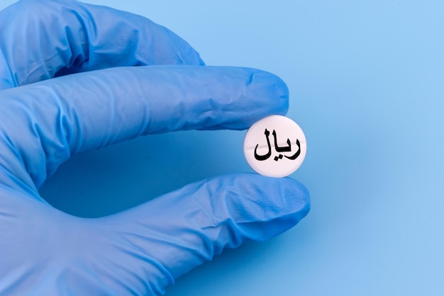 Una mano en un guante médico sostiene una píldora con un signo de rial en un fondo azul Medicina cara en Irán