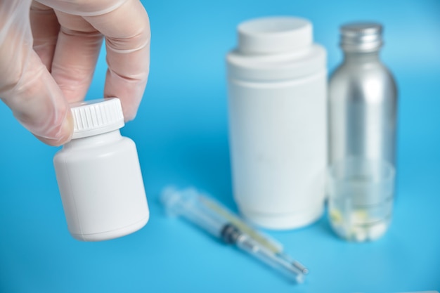 La mano en guantes sostiene un frasco blanco de medicina con en el fondo de latas médicas, píldoras en un fondo azul.