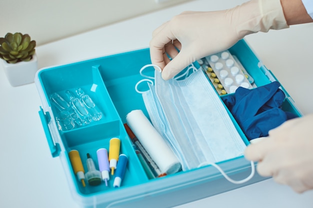 La mano en guantes toma la máscara protectora de midicina del botiquín de primeros auxilios Caja de medicina casera con artículos médicos. El virus protege el concepto.