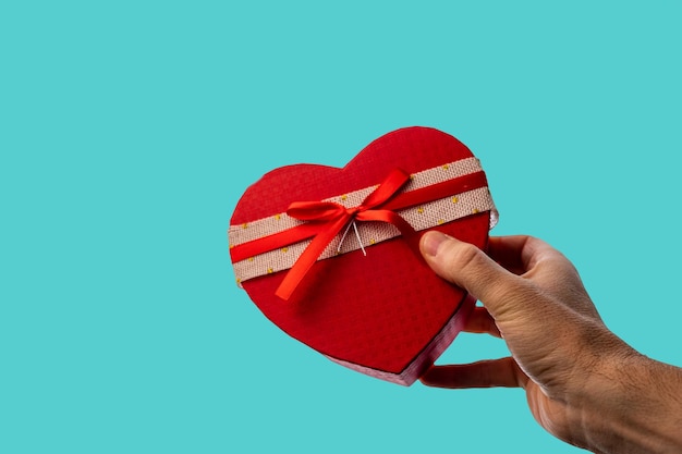 La mano del hombre dando un regalo en una caja en forma de corazón con un lazo, sobre un fondo azul.