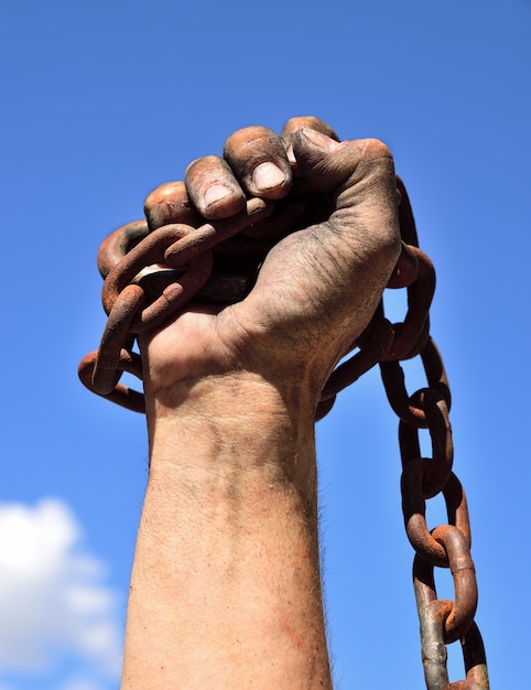 La mano del hombre envuelta en una cadena oxidada de hierro levantada contra un cielo azul