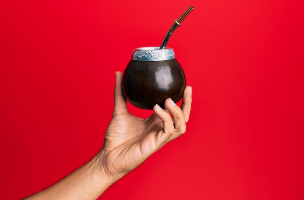 La mano de un hombre hispano sosteniendo una taza de bebida de infusión de mate sobre un fondo rojo aislado