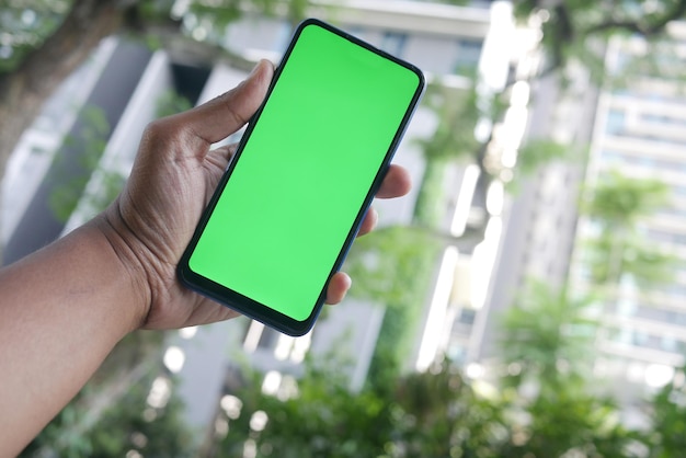 Mano de hombre joven usando un teléfono inteligente con pantalla verde contra edificios de la ciudad