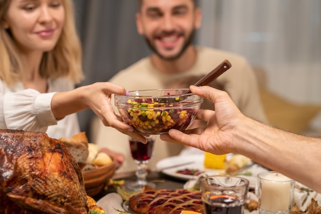 Mano de hombre maduro, pasando el tazón con ensalada casera fresca a la joven mujer rubia feliz sentada junto a la mesa festiva servida junto a su marido