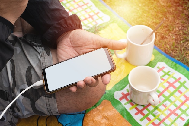 Mano del hombre mantenga smartphone sentarse en la estera con dos tazas de café vacías, al aire libre.