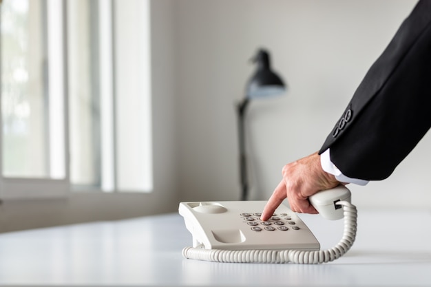 Mano de hombre de negocios marcando un número de teléfono con teléfono fijo blanco en el escritorio de oficina blanco.