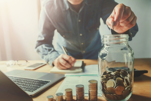 mano de hombre de negocios poniendo monedas en jarra de vidrio