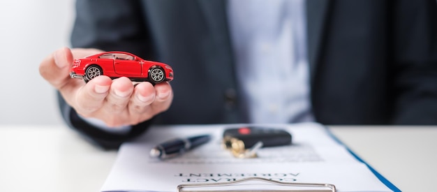 Mano de hombre de negocios que sostiene el juguete del coche rojo con la pluma sin llave del vehículo y el documento de contrato compra y venta de seguros de alquiler y conceptos de acuerdo de contrato