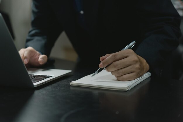 Mano de hombre de negocios sosteniendo una pluma escribiendo en un cuaderno que trabaja con una computadora portátil en una oficina moderna Escriba en el concepto de diario