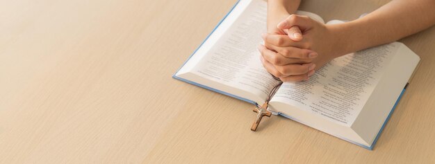 Mano de hombre orando sosteniendo la cruz en el libro de la Biblia sagrada en la mesa de madera Burgeoning