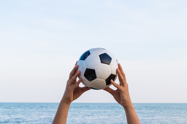 Mano del hombre que sostiene un balón de fútbol en el cielo con el mar en el fondo