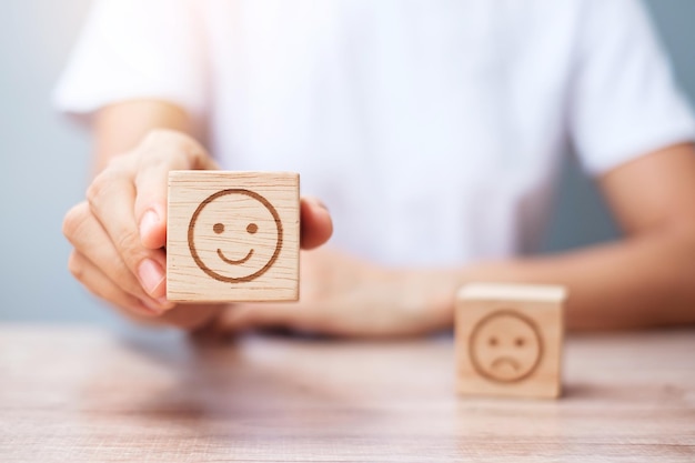Mano de hombre que sostiene el bloque de cara de emoción El cliente elige Emoticon para las revisiones de los usuarios Clasificación de calificación del servicio evaluación de satisfacción de revisión del cliente y concepto de retroalimentación