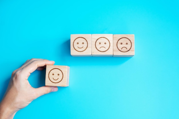 Mano de hombre que sostiene el bloque de cara de emoción El cliente elige Emoticon para las revisiones de los usuarios Clasificación de calificación del servicio evaluación de satisfacción de revisión del cliente y concepto de retroalimentación
