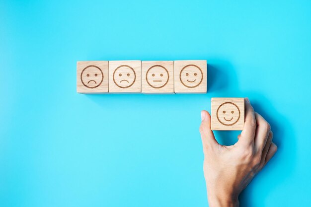 Mano de hombre que sostiene el bloque de cara de emoción El cliente elige Emoticon para las revisiones de los usuarios Clasificación de calificación del servicio evaluación de satisfacción de revisión del cliente y concepto de retroalimentación