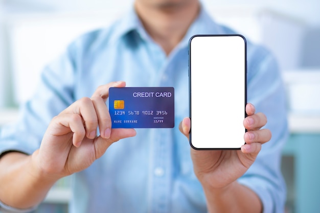 La mano del hombre que sostiene el teléfono inteligente de pantalla en blanco y la tarjeta de crédito usan camisa azul claro