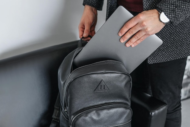 La mano del hombre saca una computadora portátil gris con una elegante mochila de cuero negro