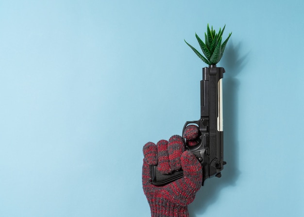 De la mano a un hombre sostenga una pistola con una planta artificial sobre fondo azul. Copie el espacio.