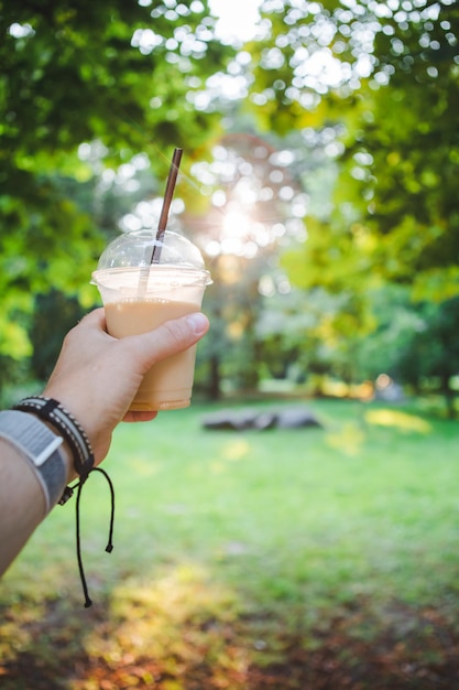 Mano de hombre sosteniendo una taza con bebida refrescante caminando por el parque público de verano