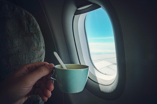 La mano de un hombre sosteniendo una taza de café frente a la ventana del avión en la que se puede ver el cielo azul y el ala del avión