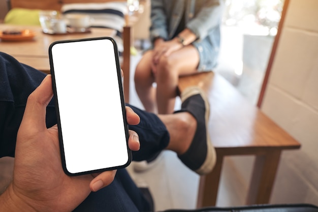La mano del hombre sosteniendo un teléfono móvil negro con pantalla en blanco con una mujer sentada en el café
