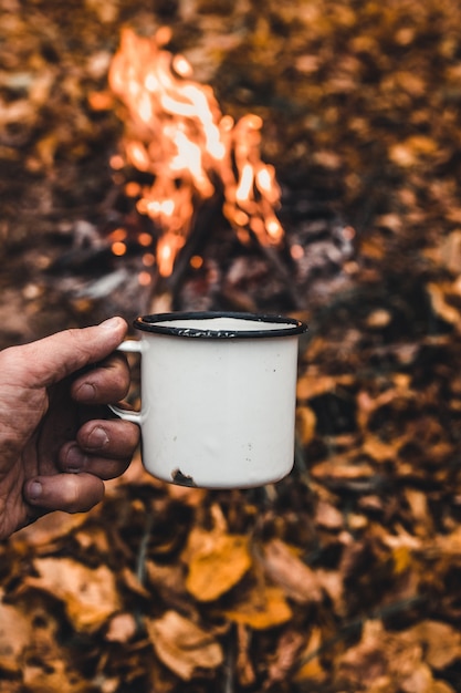 La mano del hombre sostiene una taza de café caliente en el fondo de la fogata.