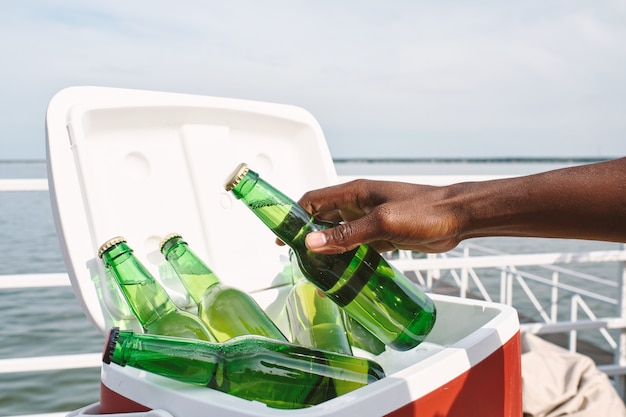 Mano del hombre tomando una botella de vidrio de sabrosa cerveza fría de una caja más fresca al asistir a la fiesta en el muelle