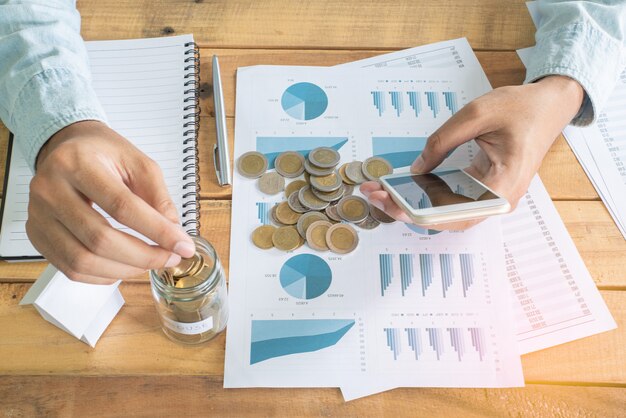 Mano del hombre usando un teléfono inteligente y poniendo monedas en el cristal en una mesa de trabajo de madera con un montón de monedas, cuaderno de notas, bolígrafo y mini casa blanca modelo - Inversión, Negocios, Finanzas y Banca